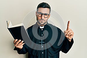 Young hispanic priest man holding bible with finger up skeptic and nervous, frowning upset because of problem
