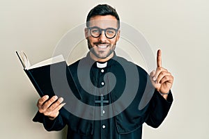 Young hispanic priest man holding bible with finger up smiling with a happy and cool smile on face