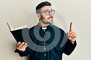 Young hispanic priest man holding bible with finger up smiling looking to the side and staring away thinking