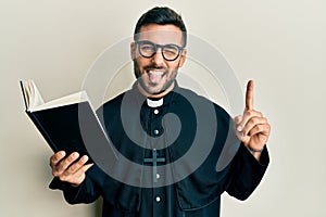 Young hispanic priest man holding bible with finger up sticking tongue out happy with funny expression