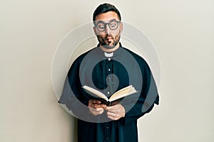 Young hispanic priest man holding bible making fish face with mouth and squinting eyes, crazy and comical