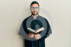 Young hispanic priest man holding bible relaxed with serious expression on face