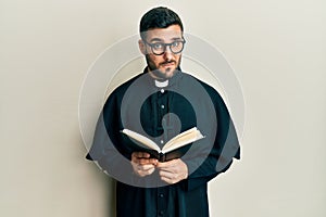 Young hispanic priest man holding bible skeptic and nervous, frowning upset because of problem