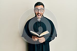 Young hispanic priest man holding bible smiling and laughing hard out loud because funny crazy joke