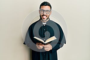 Young hispanic priest man holding bible sticking tongue out happy with funny expression