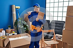 Young hispanic technician working at house renovation pointing thumb up to the side smiling happy with open mouth