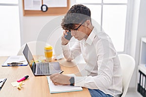 Young hispanic teenager business worker talking on smartphone writing on notebook at office