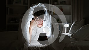 Young hispanic teenager seriously engrossed in texting on his smartphone, lying on a comfy bed under a snug blanket, in the dim