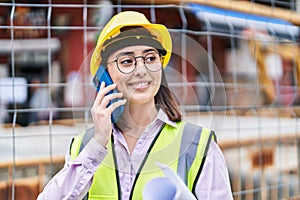 Young hispanic woman architect talking on the smartphone holding blueprints at street