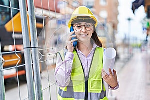 Young hispanic woman architect talking on the smartphone holding blueprints at street