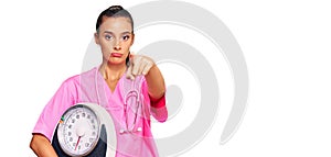Young hispanic woman as nutritionist doctor holding weighing machine pointing with finger to the camera and to you, confident