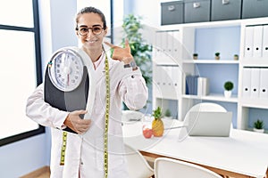 Young hispanic woman as nutritionist doctor holding weighing machine pointing finger to one self smiling happy and proud