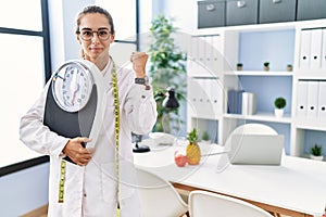 Young hispanic woman as nutritionist doctor holding weighing machine screaming proud, celebrating victory and success very excited