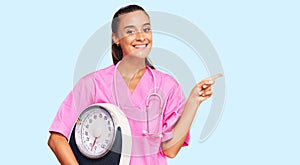 Young hispanic woman as nutritionist doctor holding weighing machine smiling happy pointing with hand and finger to the side