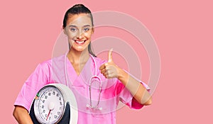 Young hispanic woman as nutritionist doctor holding weighing machine smiling happy and positive, thumb up doing excellent and