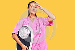 Young hispanic woman as nutritionist doctor holding weighing machine stressed and frustrated with hand on head, surprised and