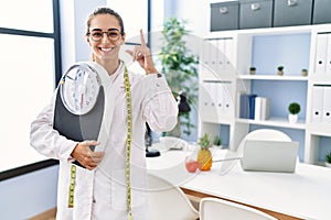 Young hispanic woman as nutritionist doctor holding weighing machine surprised with an idea or question pointing finger with happy