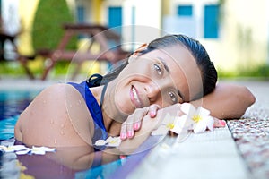 Young hispanic woman in blue dress relaxing by the swimming pool surrounded by flowers