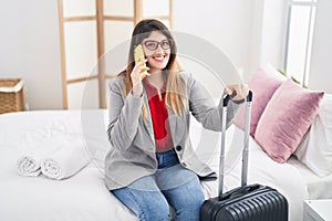 Young hispanic woman business worker talking on the smartphone sitting on bed at hotel room