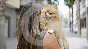 Young hispanic woman, captured from the back standing on a sunny city street, while backwards hugging her beautiful dog, embodying