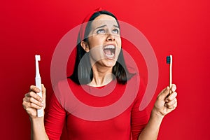 Young hispanic woman choosing electric toothbrush or normal teethbrush angry and mad screaming frustrated and furious, shouting