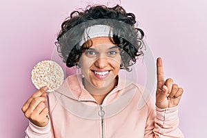 Young hispanic woman with curly hair eating healthy rice crackers smiling with an idea or question pointing finger with happy