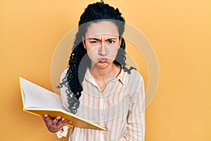 Young hispanic woman with curly hair holding book puffing cheeks with funny face