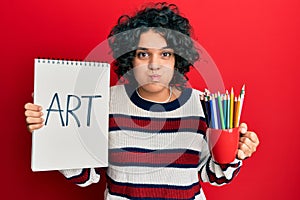 Young hispanic woman with curly hair holding canvas book and colored pencils puffing cheeks with funny face