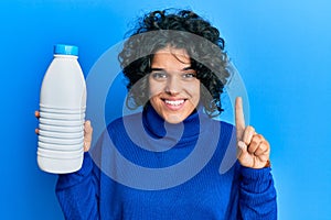 Young hispanic woman with curly hair holding liter bottle of milk smiling with an idea or question pointing finger with happy