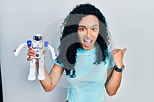 Young hispanic woman with curly hair holding robot toy pointing thumb up to the side smiling happy with open mouth