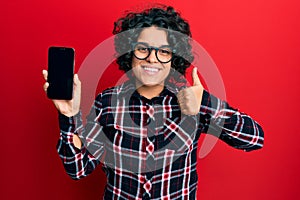 Young hispanic woman with curly hair holding smartphone showing blank screen smiling happy and positive, thumb up doing excellent