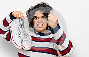 Young hispanic woman with curly hair holding white casual shoes annoyed and frustrated shouting with anger, yelling crazy with