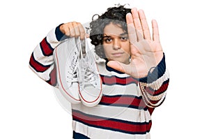 Young hispanic woman with curly hair holding white casual shoes with open hand doing stop sign with serious and confident