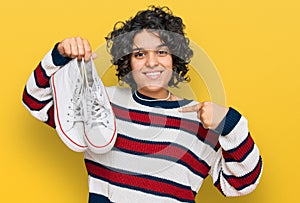 Young hispanic woman with curly hair holding white casual shoes pointing finger to one self smiling happy and proud