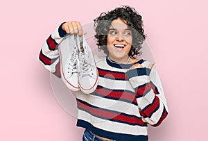 Young hispanic woman with curly hair holding white casual shoes pointing thumb up to the side smiling happy with open mouth