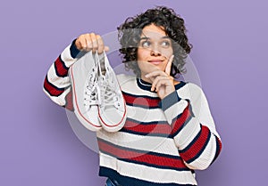 Young hispanic woman with curly hair holding white casual shoes serious face thinking about question with hand on chin, thoughtful