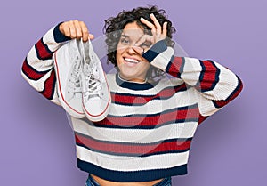Young hispanic woman with curly hair holding white casual shoes smiling happy doing ok sign with hand on eye looking through