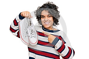 Young hispanic woman with curly hair holding white casual shoes smiling happy pointing with hand and finger