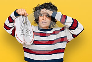 Young hispanic woman with curly hair holding white casual shoes stressed and frustrated with hand on head, surprised and angry