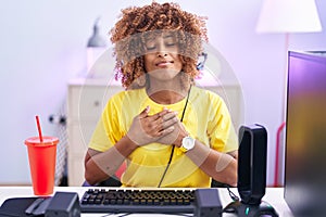 Young hispanic woman with curly hair playing video games wearing headphones smiling with hands on chest with closed eyes and