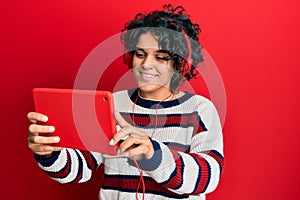 Young hispanic woman with curly hair using touchpad wearing headphones looking positive and happy standing and smiling with a