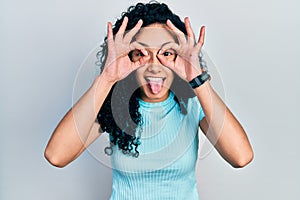 Young hispanic woman with curly hair wearing casual blue t shirt doing ok gesture like binoculars sticking tongue out, eyes