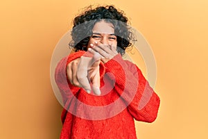 Young hispanic woman with curly hair wearing casual winter sweater laughing at you, pointing finger to the camera with hand over