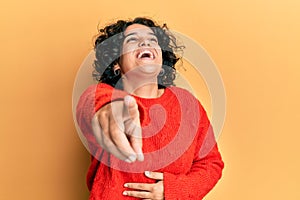 Young hispanic woman with curly hair wearing casual winter sweater laughing at you, pointing finger to the camera with hand over