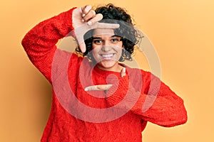 Young hispanic woman with curly hair wearing casual winter sweater smiling making frame with hands and fingers with happy face