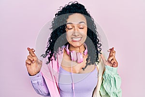 Young hispanic woman with curly hair wearing gym clothes and using headphones gesturing finger crossed smiling with hope and eyes