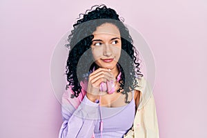 Young hispanic woman with curly hair wearing gym clothes and using headphones with hand on chin thinking about question, pensive