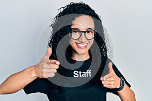 Young hispanic woman with curly hair wearing staff t shirt pointing fingers to camera with happy and funny face