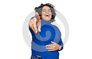 Young hispanic woman with curly hair wearing turtleneck sweater laughing at you, pointing finger to the camera with hand over