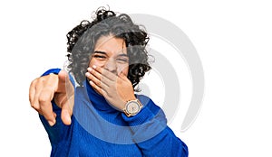 Young hispanic woman with curly hair wearing turtleneck sweater laughing at you, pointing finger to the camera with hand over